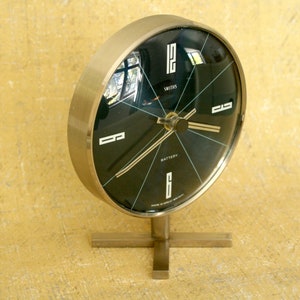Puede incluir: Un reloj de escritorio redondo Smiths que funciona con pilas, con un marco de tono plateado metálico y una esfera negra. El reloj presenta números blancos en las posiciones de las 12, 3, 6 y 9, manecillas plateadas finas y el texto "SMITHS" y "MADE IN GREAT BRITAIN."