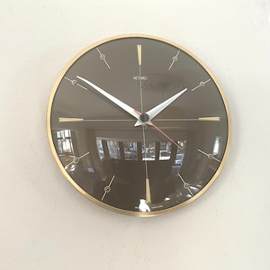 May include: A round wall clock with a gold-tone metal frame and a dark brown glass face. The clock features white hour and minute hands, a thin red second hand, and gold-tone markers. The text METAMEC is printed at the top, and MADE IN ENGLAND at the bottom.