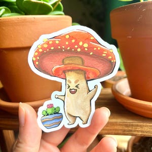 Pode incluir: Um adesivo recortado apresentando um personagem de cogumelo antropomórfico bege com um grande chapéu vermelho coberto de manchas amarelas. O personagem tem uma expressão alegre e segura um pequeno vaso azul com um cacto verde e flor rosa.
