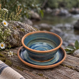 Puede incluir: Un juego de cuenco y plato de cerámica con un esmalte de remolinos azules y verde azulado, con bordes de arcilla color terracota. Las piezas tienen un aspecto redondeado y hecho a mano, con anillos concéntricos visibles en el interior.
