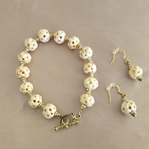 May include: A white bracelet with gold accents and matching earrings. The bracelet is made of round beads with a carved floral design. The earrings are dangle style with a round bead at the bottom.