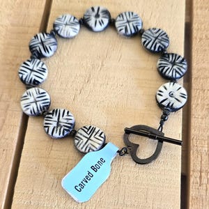 May include: A black and white carved bone bracelet with a heart-shaped clasp. The tag reads "Carved Bone".