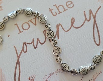 Könnte beinhalten: Silberfarbenes Armband mit spiralförmigen Charms. Das Armband hat einen Knebelverschluss und einen floralen Charm. Im Hintergrund steht "love the journey".
