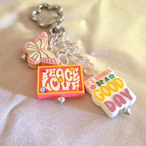 Puede incluir: Un llavero de tono plateado con una anilla en forma de flor y tres dijes: una mariposa rosa y blanca, un dije rectangular naranja con el texto PEACE LOVE y un dije blanco con el texto IT'S GONNA BE A GOOD DAY en amarillo y rosa.