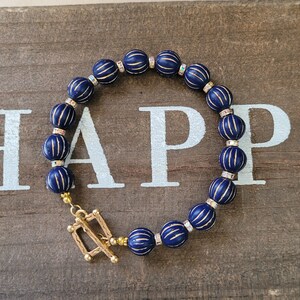 May include: A navy blue beaded bracelet with gold detailing. The bracelet has round, ribbed beads with gold stripes, interspersed with small, clear crystal beads. A gold-coloured clasp secures the bracelet. The word "HAPPY" is visible in the background.