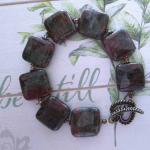 May include: A bracelet featuring square, polished stones in shades of burgundy and green. The stones are strung together with small, iridescent beads. The bracelet is secured with a decorative, antique-style toggle clasp. The bracelet is a unique piece of jewelry.