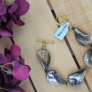 May include: A bracelet made of iridescent, teardrop-shaped Mother of Pearl beads. The beads are strung together with small gold-colored beads and a gold-tone clasp. A tag that reads "Mother of Pearl" is attached to the bracelet. The bracelet is displayed on a wooden surface.