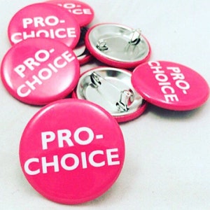 May include: Several bright pink, circular badges with the text "PRO-CHOICE" in white lettering. Some badges are face up, while others reveal the silver pin backs. The badges are arranged on a white background.