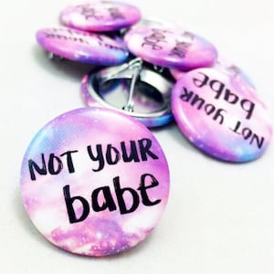 Könnte beinhalten: Ein Satz runder, rosa und lilafarbener Buttons im Galaxie-Design mit dem in Schwarz gedruckten Text "Not your babe".