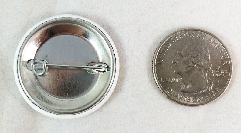 May include: The back of a white button with a silver pin and a silver backing. The button is about the size of a US quarter.