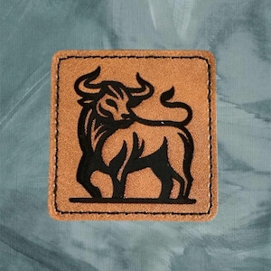 May include: A brown leather patch with a black silhouette of a bull. The bull is facing the right side of the image and has its head turned to the left. The patch has a stitched border.