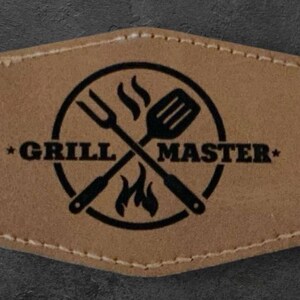May include: A brown leather patch with a black circle containing a crossed spatula and fork with flames. The text "*GRILL MASTER*" is printed around the circle.