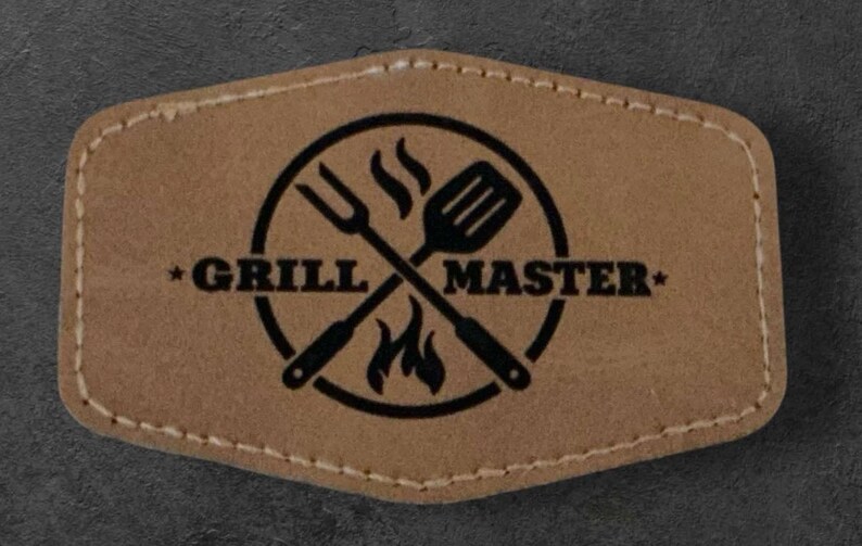 May include: A brown leather patch with a black circle containing a crossed spatula and fork with flames. The text "*GRILL MASTER*" is printed around the circle.
