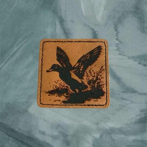 May include: A brown leather patch with a black silhouette of a duck in flight. The duck is in the center of the patch and is facing to the right. The patch has a stitched border.