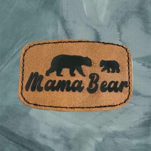 May include: A brown leather patch with a black silhouette of a mama bear and a baby bear. The text "Mama Bear" is written in black lettering on the patch.