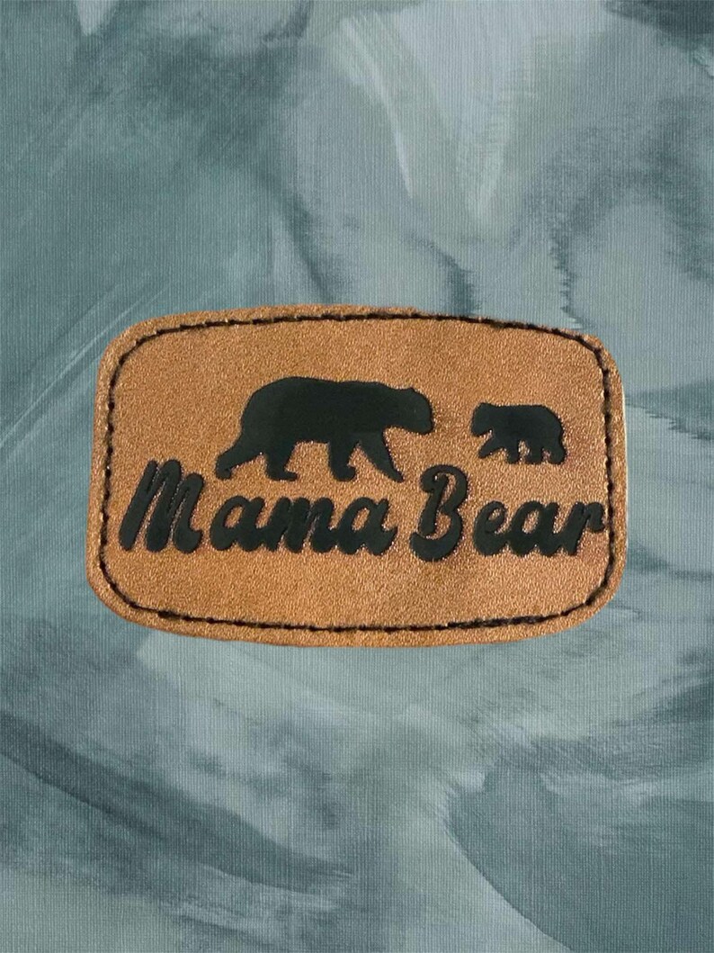May include: A brown leather patch with a black silhouette of a mama bear and a baby bear. The text "Mama Bear" is written in black lettering on the patch.