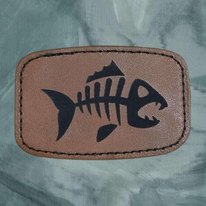 May include: A brown leather patch with a black fish skeleton design. The fish has a large mouth and a fin on its back.