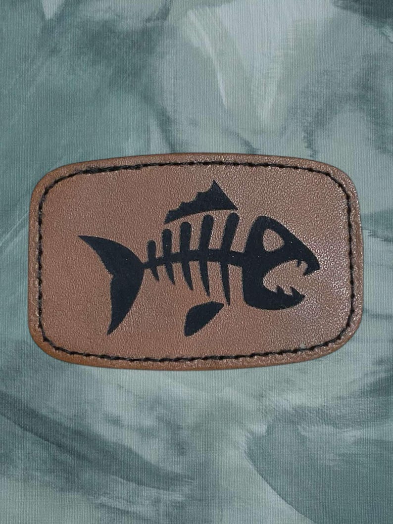 K&ouml;nnte beinhalten: Ein braunes Leder-Patch mit einem schwarzen Fischskelett-Design. Der Fisch hat einen gro&szlig;en Mund und eine R&uuml;ckenflosse.