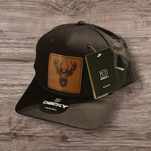 May include: Black and brown trucker hat with a brown leather patch featuring a deer silhouette. The hat has a black mesh back and a black brim. The hat has a "DECKY ORIGINAL" label on the front.
