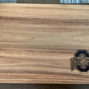 May include: Wooden cutting board with a carved border and the Ohio State logo. The board is rectangular and made of light brown wood. The logo is black and white.