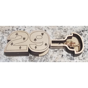May include: A light-coloured wooden box shaped like the number 29, featuring engraved card suit symbols including clubs, hearts, and diamonds. Beside it lies a matching open wooden container shaped like a guitar, holding several small metal pins.