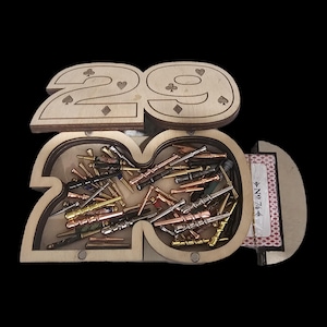 May include: A light-colored wooden box shaped like the number 29, featuring engraved card suit symbols on the lid. The open container holds a collection of small, metallic, wand-like pegs in various finishes, with a deck of No. 74 playing cards.