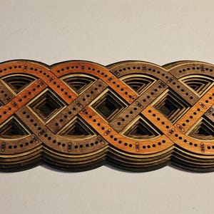 May include: A wooden cribbage board with a Celtic knot design. The board is made of layered wood in shades of brown and orange, with small holes for pegs. The design is intricate and geometric.