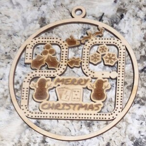 May include: Wooden Christmas ornament with a circular design. The ornament features the text "MERRY CHRISTMAS" and includes images of snowmen, holly, snowflakes, and a reindeer pulling a sleigh. The ornament has a small hole at the top for hanging.
