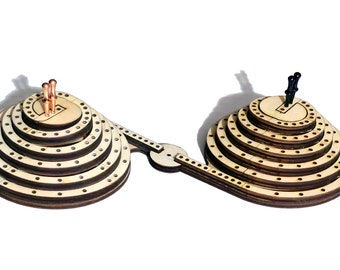 May include: A wooden game with two circular, layered platforms connected by a track. Each platform has a central hole and a series of concentric circles with holes around the edge. There are three copper pegs on one platform and two black pegs on the other.