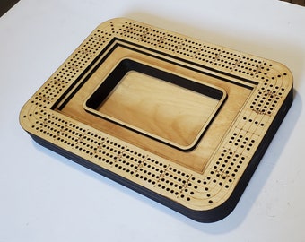 May include: A wooden cribbage board with a black border and a removable playing surface. The board has a traditional design with numbered holes and a scoring track.