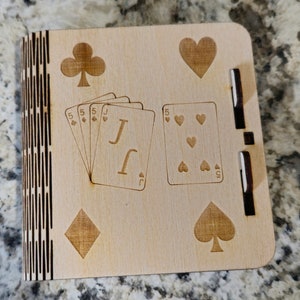 May include: A wooden box with a hinged lid, featuring a laser-engraved design of playing cards. The box has a black and white striped pattern on the side and a black border around the lid. The lid features a jack of clubs, a five of hearts, a diamond, and a spade.