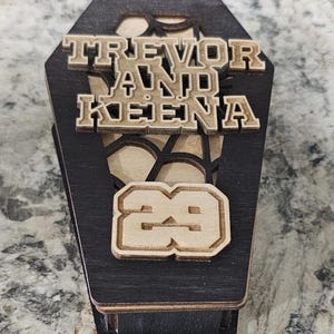 May include: A black wooden coffin-shaped box with a spiderweb design. The box has the names "Trevor and Keiena" and the number "29" engraved on it.