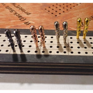 May include: A close-up of a cribbage board with metal pegs in various colors. The pegs include black, copper, silver, and gold. The board is made of wood with a light-colored surface and a dark border. The text "If (Jack.Suit == printed" is visible.