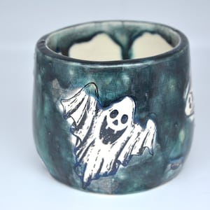 May include: A ceramic mug featuring a dark teal and black mottled glaze with a hand-drawn white ghost character on the exterior. The ghost has a wide, smiling mouth and hollow eyes. The interior of the mug is a plain, light cream colour.