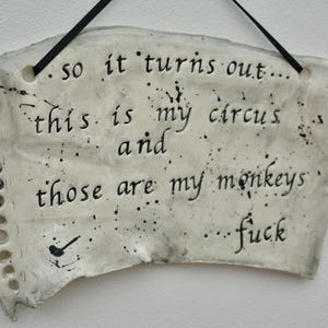 May include: A white ceramic wall plaque shaped like a torn piece of notebook paper with black ink splatters. The text reads: so it turns out... this is my circus and those are my monkeys...fuck. A black ribbon is threaded through two top holes.