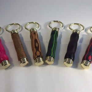 May include: Six wooden keychain bottle openers with different wood grain patterns and colors. The keychains are gold colored metal.