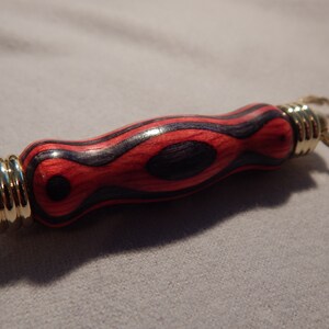 May include: A wooden keychain with a red and black wood grain pattern and a gold metal ring. The keychain is shaped like a small cylinder.