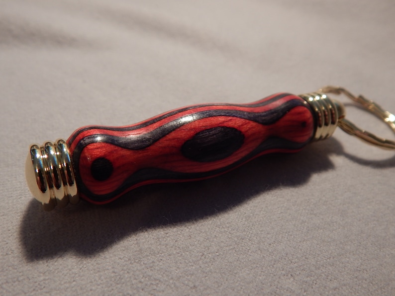May include: A wooden keychain with a red and black wood grain pattern and a gold metal ring. The keychain is shaped like a small cylinder.