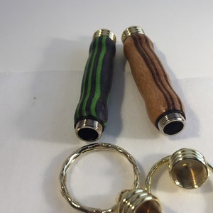 May include: Two wooden keychains with different wood grain patterns. One is black and green, the other is brown and white. Both have gold metal caps on the ends.