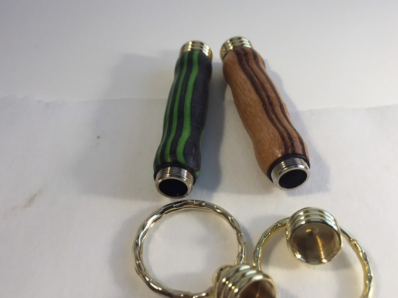 May include: Two wooden keychains with different wood grain patterns. One is black and green, the other is brown and white. Both have gold metal caps on the ends.