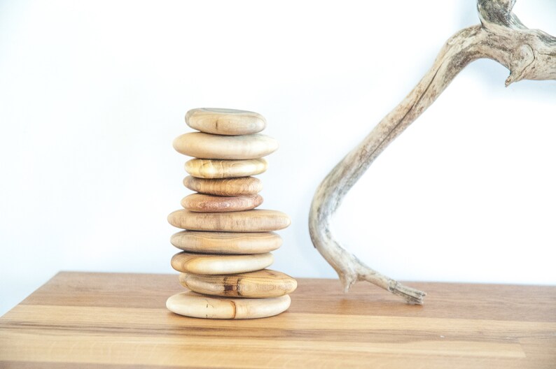 May include: A stack of smooth, light brown wooden discs, resembling a zen rock garden, sits on a wooden surface. A light brown driftwood branch is in the background.