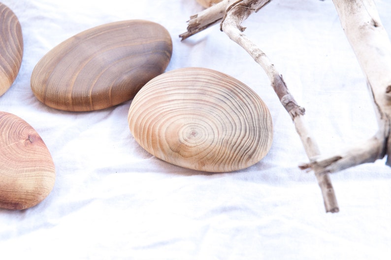 May include: Four wooden coasters with a natural wood finish. The coasters are round and oval shaped and have a smooth surface. The coasters are arranged on a white fabric background.