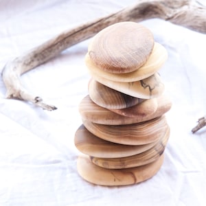 May include: A stack of seven wooden coasters, each with a unique grain pattern and shape, arranged in a pyramid on a white fabric background.