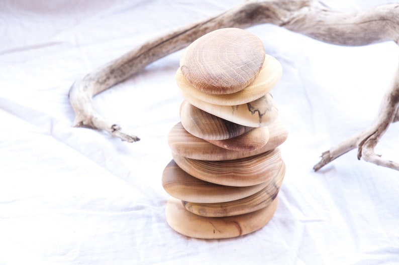 May include: A stack of seven wooden coasters, each with a unique grain pattern and shape, arranged in a pyramid on a white fabric background.