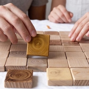May include: A set of square wooden tiles arranged in a grid pattern on a white surface. Each tile features a distinct natural wood grain pattern, with some displaying circular tree ring designs in shades of light tan, medium brown, and dark brown.