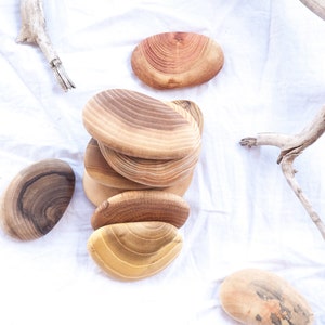 May include: A stack of six natural wood oval-shaped coasters with a light brown wood grain pattern. The coasters are arranged in a pile on a white fabric surface.
