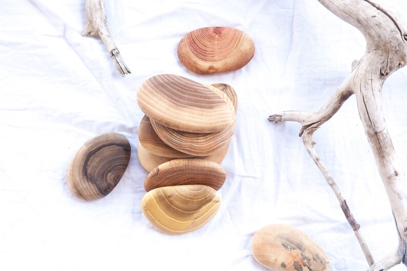 May include: A stack of six natural wood oval-shaped coasters with a light brown wood grain pattern. The coasters are arranged in a pile on a white fabric surface.