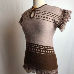 Peut inclure: Un pull en tricot marron et beige avec un motif géométrique et une bordure en franges. Le pull a un col en forme de trou de serrure avec un bouton en bois.