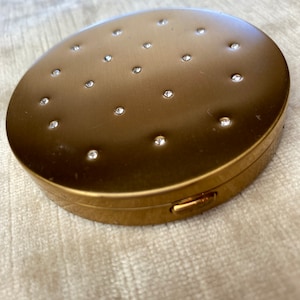Peut inclure: Un compact rond doré avec un motif décoratif de petits strass.