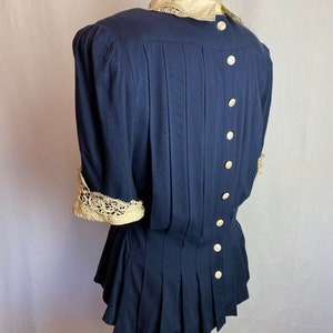 May include: A navy blue blouse with a white lace collar and cuffs. The blouse has a fitted bodice and a flared skirt. It features a row of white buttons down the back.